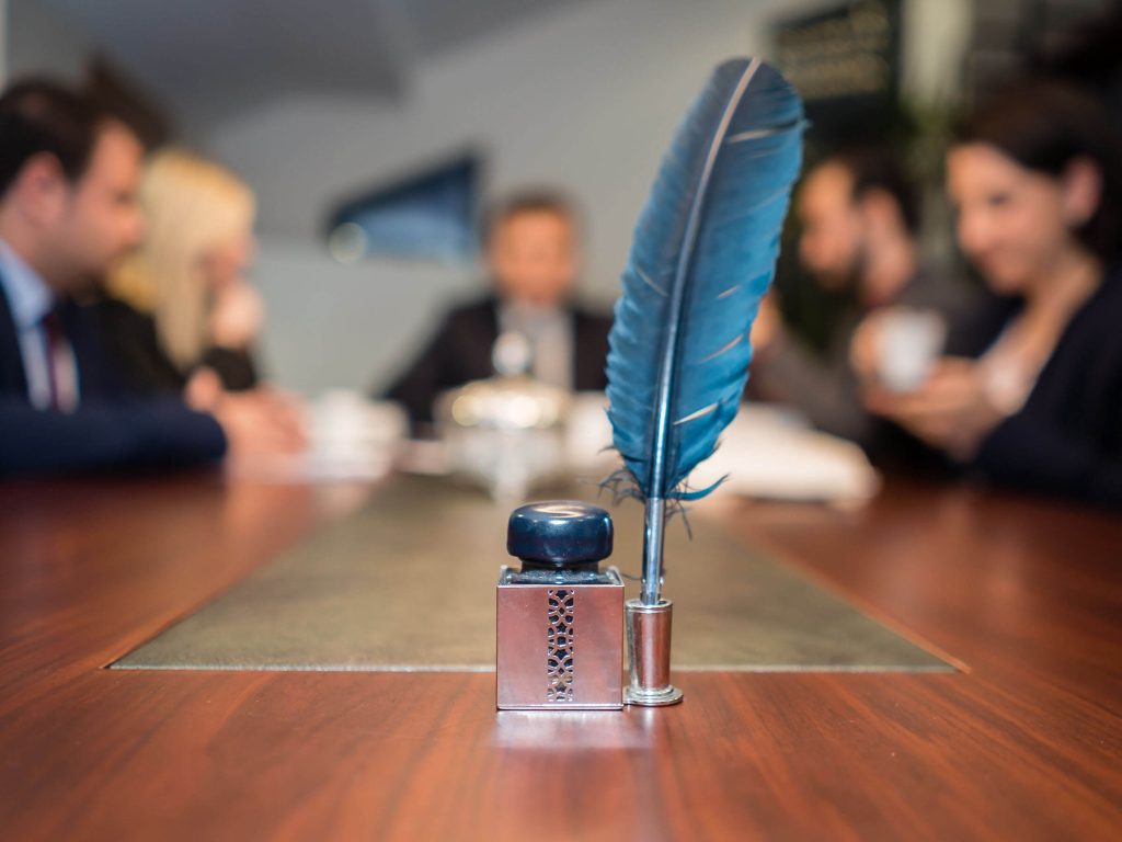 Tintero antiguo con pluma estilográfica sobre una mesa de reuniones, al fondo un grupo de profesionales discuten un caso legal.